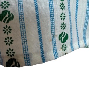 May include: Close-up of white fabric with blue vertical stripes and green floral and cloud-like patterns. The fabric has a textured appearance, possibly part of a larger textile item.