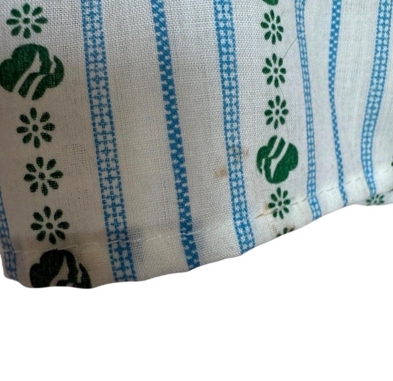 May include: Close-up of white fabric with blue vertical stripes and green floral and cloud-like patterns. The fabric has a textured appearance, possibly part of a larger textile item.