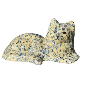 May include: A ceramic cat figurine in a reclining pose. The cat is covered in a floral pattern of blue flowers on a cream-colored background. The cat has black eyes and a black nose.