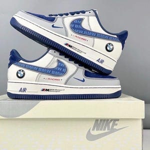 以下が含まれることがあります： BMWのブランドが入った、白、グレー、ネイビーブルーのナイキエアフォース1スニーカー。靴にはBMWのロゴが側面とヒールにあり、「BMW Motorrad Motorsport」の文字が入っています。スニーカーはナイキの箱に展示されています。