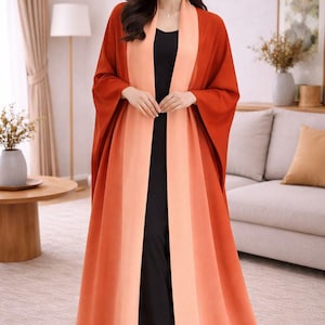 Pode incluir: Uma mulher usando um robe longo e aberto em tons de laranja e pêssego sobre um traje preto. O robe tem um design de blocos de cores com listras verticais. O fundo inclui uma sala de estar com um sofá, mesa e lâmpada.