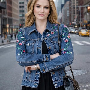 Könnte beinhalten: Hellblaue Jeansjacke mit bunten Blumenstickereien. Die Jacke hat ein klassisches Design mit Knopfverschlüssen und wird über einem schwarzen Oberteil getragen. Eine schwarze, gesteppte Handtasche mit Kettenriemen ist ebenfalls zu sehen.