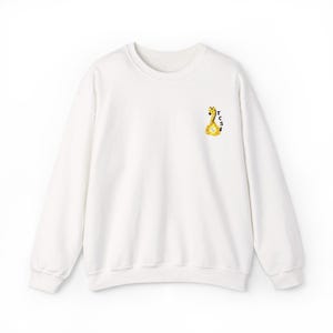 May include: A white long-sleeve crewneck sweatshirt featuring a small embroidered graphic on the upper left chest. The graphic depicts a yellow and brown giraffe with the vertical black text TCS next to it, positioned on the right side of the giraffe.