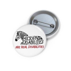 May include: White button with a black and white zebra design. The text "HIDDEN DISABILITIES" is incorporated into the zebra's body, with "ARE REAL DISABILITIES" in red below. A second button is visible in the background.