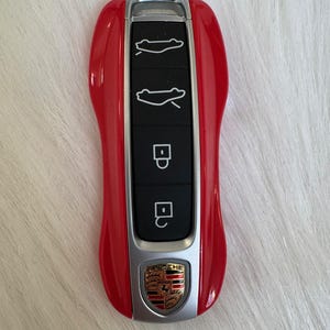 May include: A red car key with silver accents and a Porsche emblem. The key has buttons for car functions, including opening the trunk and locking the doors. The key is a sleek, modern design.