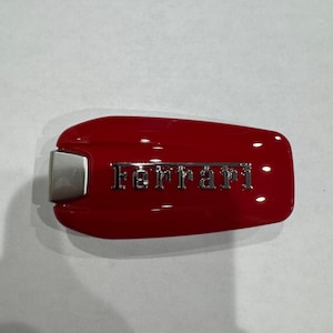 Pode incluir: Chave do carro Ferrari vermelha com o nome da marca em letras prateadas. A chave tem uma forma retangular arredondada com uma extremidade de metal prateado.