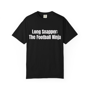 May include: Black t-shirt with the text "Long Snapper: The Football Ninja" in white block letters. The shirt is a basic crew neck style, suitable for casual wear. The text is centered on the front of the shirt.