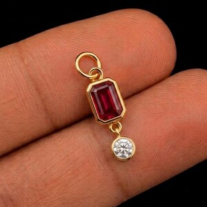 May include: A gold pendant featuring a rectangular emerald-cut red gemstone set in a bezel, with a small round white diamond suspended below it. The pendant includes a circular jump ring at the top for attachment to a chain or necklace.