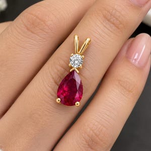 May include: A gold pendant featuring a pear-shaped red gemstone suspended below a single round white gemstone. The pendant is held in a gold setting with a small bail at the top for attachment to a chain, showcasing a polished metal finish.