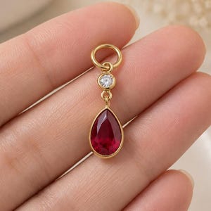 Puede incluir: Un colgante de oro con una piedra preciosa roja en forma de lágrima suspendida de una pequeña anilla circular, con una piedra redonda transparente engastada entre la anilla y la lágrima roja. El engaste es de metal dorado pulido.