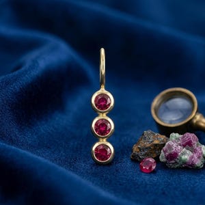 Op de afbeelding: Een gouden oorbel met drie ronde, rode edelstenen. De oorbel wordt getoond op een blauw fluwelen oppervlak, met een vergrootglas, een robijn en wat stenen op de achtergrond. De edelstenen zijn gezet in gouden zettingen.