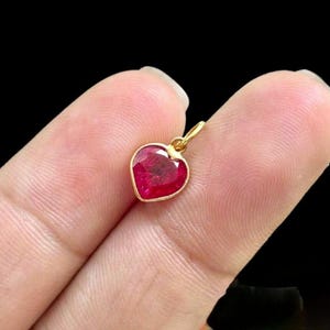 May include: A heart-shaped pendant with a vibrant red gemstone set in a gold frame. The pendant is suspended from a small gold loop, perfect for a necklace or bracelet. The gemstone has a polished, reflective surface.