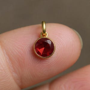 May include: A small, round pendant with a faceted, deep red gemstone set in a gold-colored bezel. The pendant has a small loop at the top for attaching to a chain or bracelet. The gemstone is approximately 0.25 inches in diameter.