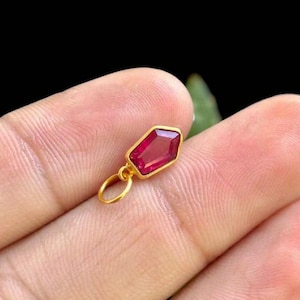 Op de afbeelding: Een kleine, goudkleurige hanger met een gefacetteerde, robijnrode edelsteen. De edelsteen is gezet in een gouden bezel en heeft een unieke, geometrische vorm. De hanger heeft een kleine lus.