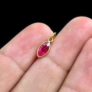 Op de afbeelding: Een gouden hanger met een marquise-geslepen, dieprode edelsteen. De edelsteen is gezet in een gouden bezel en de hanger heeft een kleine lus voor een ketting. Dit is waarschijnlijk een sieraad.