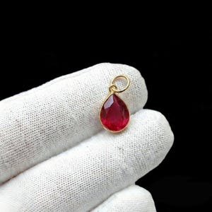 Puede incluir: Un colgante en forma de lágrima con una gema facetada de color rojo intenso engastada en un bisel dorado. El colgante tiene un pequeño lazo en la parte superior para una cadena. El fondo es negro.