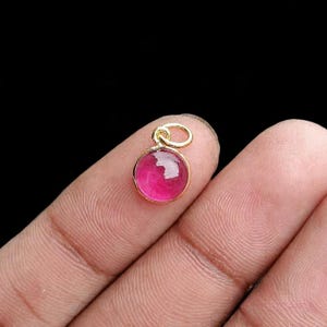 Puede incluir: Un pequeño colgante redondo con una gema rosa vibrante engastada en un marco de metal dorado. El colgante tiene un lazo en la parte superior para una cadena. La gema pulida refleja la luz, creando una apariencia brillante. El fondo es negro.