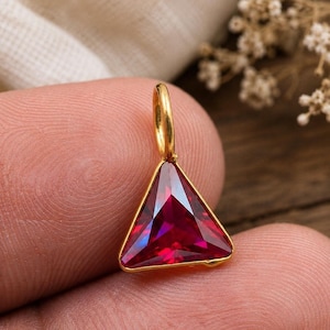 May include: A triangular pendant featuring a faceted deep red gemstone set in a polished gold-toned metal frame. The pendant includes a small circular gold-toned bail at the top for attachment to a chain, showcasing a vibrant, reflective surface.