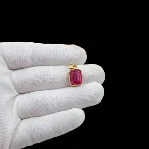 May include: A rectangular, ruby-coloured gemstone pendant with a gold-coloured setting. The pendant is suspended from a small, gold-coloured loop. The gemstone is a vibrant pinkish-red colour. The background is black.