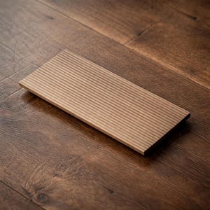 以下が含まれることがあります： 溝のある表面を持つ、長方形の明るい茶色の木製板。 板は濃い茶色の木の床に斜めに配置されており、色と質感のコントラストを生み出しています。 溝は互いに平行に走っています。