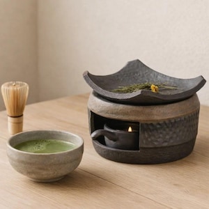 May include: A Japanese tea ceremony set. It includes a matcha bowl with green tea, a bamboo whisk, and a ceramic incense burner with a candle. The incense burner has a dark grey, textured design and a decorative top.