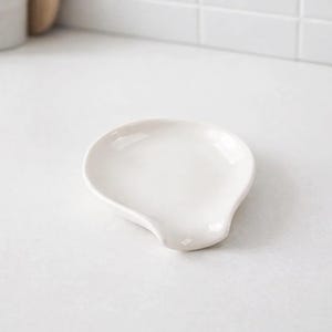 Pode incluir: Um descanso de colher de cerâmica branca com um design curvo. O descanso de colher tem formato oval com uma pequena reentrância curva em um lado. A superfície é lisa e brilhante, refletindo a luz. O item de cozinha foi projetado para segurar colheres e outros utensílios de cozinha.