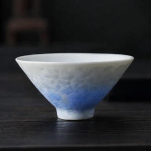 May include: A white ceramic tea bowl with a flared shape and a textured, crackled surface. The bowl transitions from white at the rim to a gradient of blue towards the base. The bowl is designed for tea drinking.