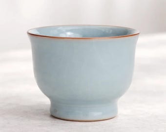 May include: A light blue ceramic teacup with a brown rim. The cup has a rounded body and a small, sturdy base. The teacup is likely used for serving tea or other hot beverages.
