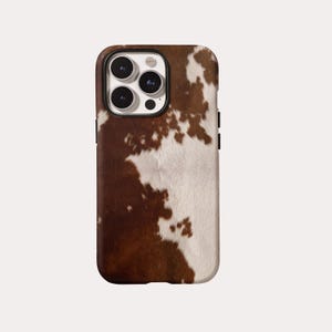 May include: A smartphone case featuring a brown and white cowhide print pattern. The case has a rectangular shape with rounded corners and a cutout at the top left corner for a triple-lens camera system, showing three black circular lenses.