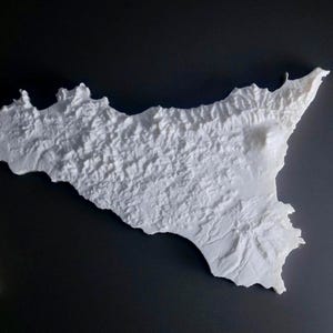 May include: A white, 3D-printed relief map of Sicily, Italy, with detailed topographical features. The map is set against a dark gray background, highlighting the island's shape and mountainous terrain. A book is partially visible in the lower left corner.