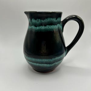 May include: A handmade ceramic jug with a black base and a teal and black glazed exterior. The jug has a curved handle and a spout for pouring. The pottery is a unique piece of functional art.