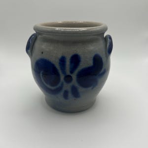 Puede incluir: Una pequeña olla de cerámica gris con dos asas pequeñas y un diseño floral azul oscuro. La olla tiene forma redondeada y un borde ligeramente ensanchado. El diseño azul es simétrico y cubre parte de la superficie de la olla.