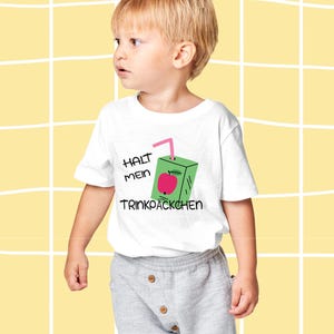 Può includere: Un bambino piccolo indossa una maglietta bianca con una grafica di scatola di succo verde e il testo tedesco "HALT MEIN TRINKPÄCKCHEN". Il bambino indossa anche pantaloni grigi e scarpe nere. Lo sfondo è una griglia gialla.