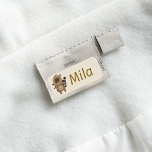Peut inclure: Une petite étiquette rectangulaire avec le nom "Mila" en lettres dorées et une illustration de raton laveur de dessin animé. L'étiquette est fixée à un tissu de couleur claire. L'étiquette mesure 3 cm x 1,3 cm.