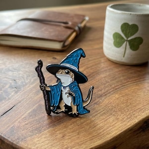 Może przedstawiać: Kolorowa emaliowana przypinka przedstawiająca jaszczurkę ubraną w strój czarodzieja, w niebieskim kapeluszu i szacie, trzymającą laskę. Przypinka znajduje się na drewnianej powierzchni, z skórzanym notatnikiem i kubkiem z koniczyną w tle.