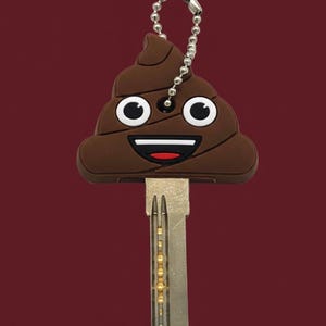 May include: A novelty key with a brown, smiling poop emoji design. The key has a silver-colored metal blade and a small chain with a clasp for attaching to a keychain. The background is a solid maroon color.