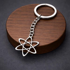 May include: Silver-colored keychain featuring a starburst design pendant. The pendant is connected to a chain and a round key ring. The keychain is displayed on a dark wooden surface against a dark background. The starburst design has six points.