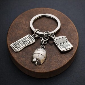 May include: Silver-colored keychain featuring a keyboard, computer mouse with the word "love", and laptop charms. The charms are attached to a silver ring. The keychain rests on a round, dark brown wooden surface.