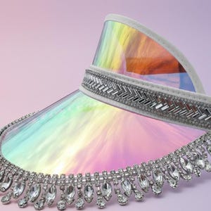 Puede incluir: Una visera elegante con un escudo transparente e iridiscente. La visera presenta una diadema blanca con un diseño geométrico plateado y un borde inferior adornado con cristales transparentes colgantes. La visera se encuentra sobre un fondo púrpura suave.