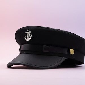 May include: A black sailor hat with a silver anchor emblem. The hat features a black leather visor, a black band, and a gold button on the side. The hat is set against a soft pink and purple background.