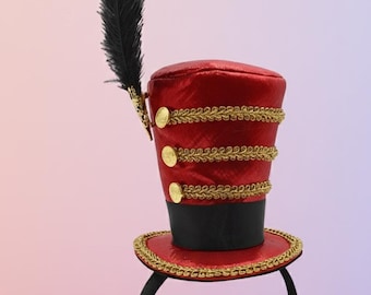 Peut inclure: Un serre-tête chapeau haut de forme rouge et noir avec une garniture dorée et une plume noire. Le chapeau a trois boutons dorés et est fixé à un serre-tête noir. Accessoire de costume.