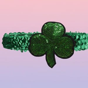 以下が含まれることがあります： 大きなクローバーのアップリケが付いた、緑色のスパンコールヘッドバンド。ヘッドバンドは、小さく光沢のある緑色のスパンコールで覆われています。クローバーも緑色でスパンコール付き、黒い縁取りです。背景はピンクと紫のグラデーションです。