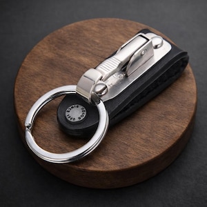 May include: A black leather key holder with a silver metal clasp and key ring. The key holder is on a round, dark brown wooden coaster. The key ring has a small, round black leather piece with the text "SINCE 1888".