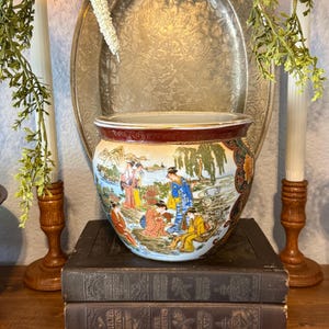 May include: A round ceramic planter featuring a colorful hand-painted scene of figures in traditional attire within a landscape. The bowl has a wide opening with a gold-trimmed rim and a deep red decorative border pattern around the top edge.