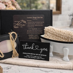 May include: A black gift box set with a soap bar, a loofah in a miniature white bathtub, and a thank you card. The set includes a soap dish and a lavender and cream soap bar. The text on the card says "thank you".