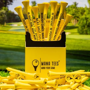 May include: A yellow box of Mana Tees golf tees with inspirational phrases like "Think Less, Enjoy More" and "Be Present, Be Positive." The tees are scattered on green grass, with a golf course in the background.