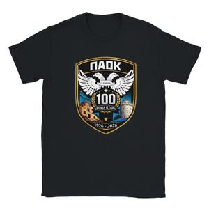 May include: A black short-sleeved t-shirt featuring a central shield-shaped graphic. The design includes a white double-headed eagle, the text "ΠΑΟΚ", "100", "ΧΡΟΝΙΑ ΙΣΤΟΡΙΑ", and "1926 - 2026", along with illustrations of a stone building and a white tower.