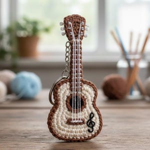 Könnte beinhalten: Ein handgefertigter Gitarren-Schlüsselanhänger aus Häkelarbeit in Braun und Creme. Die Miniatur-Gitarre hat ein Schallloch, Saiten und ein Notenschlüssel-Symbol. Der Schlüsselanhänger ist an einer silbernen Kette befestigt. Im Hintergrund sind Wollknäuel und ein Fenster zu sehen.