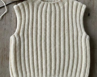 Könnte beinhalten: Cremefarbene, gerippte Strickweste. Das ärmellose Kleidungsstück hat einen Rundhalsausschnitt und ein vertikal geripptes Design. Ein Knäuel Wolle und eine Stricknadel befinden sich im Hintergrund. Die Weste ist wahrscheinlich handgefertigt.