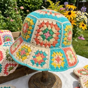 Può includere: Un cappello a secchiello all'uncinetto colorato con un design patchwork. Il cappello presenta quadrati nei toni del blu, crema, giallo, arancione e rosa. L'orlo e la fascia superiore sono turchesi. Il cappello è esposto su un supporto di legno.